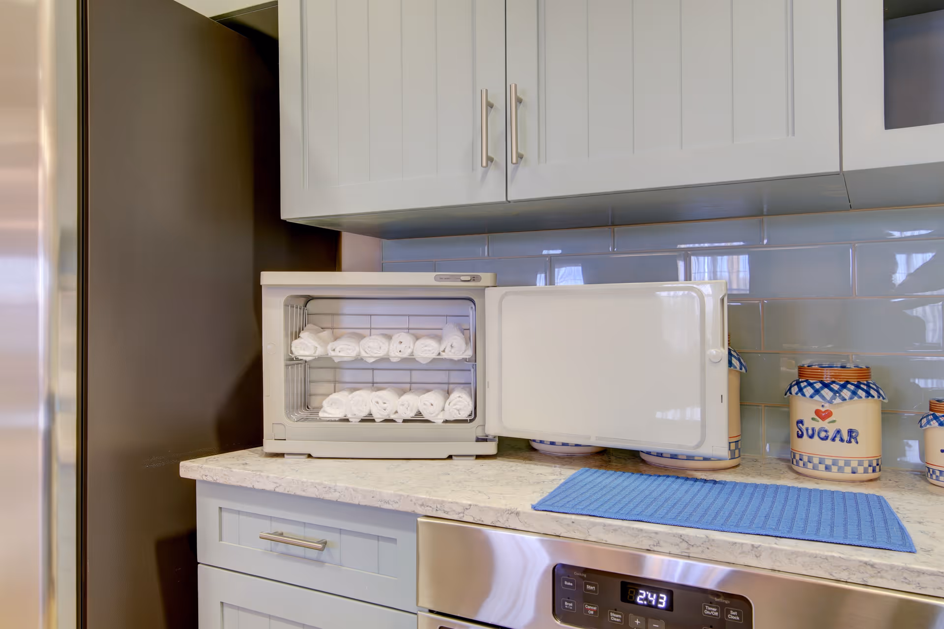 A kitchen countertop with a small white towel warmer containing rolled white towels. Above the countertop are white cabinets with silver handles. To the right on the counter are ceramic containers labeled 'Sugar' and other kitchen essentials. A blue mat is placed on the countertop next to the towel warmer. Below the counter is a stainless steel oven with a digital clock displaying 2:43.