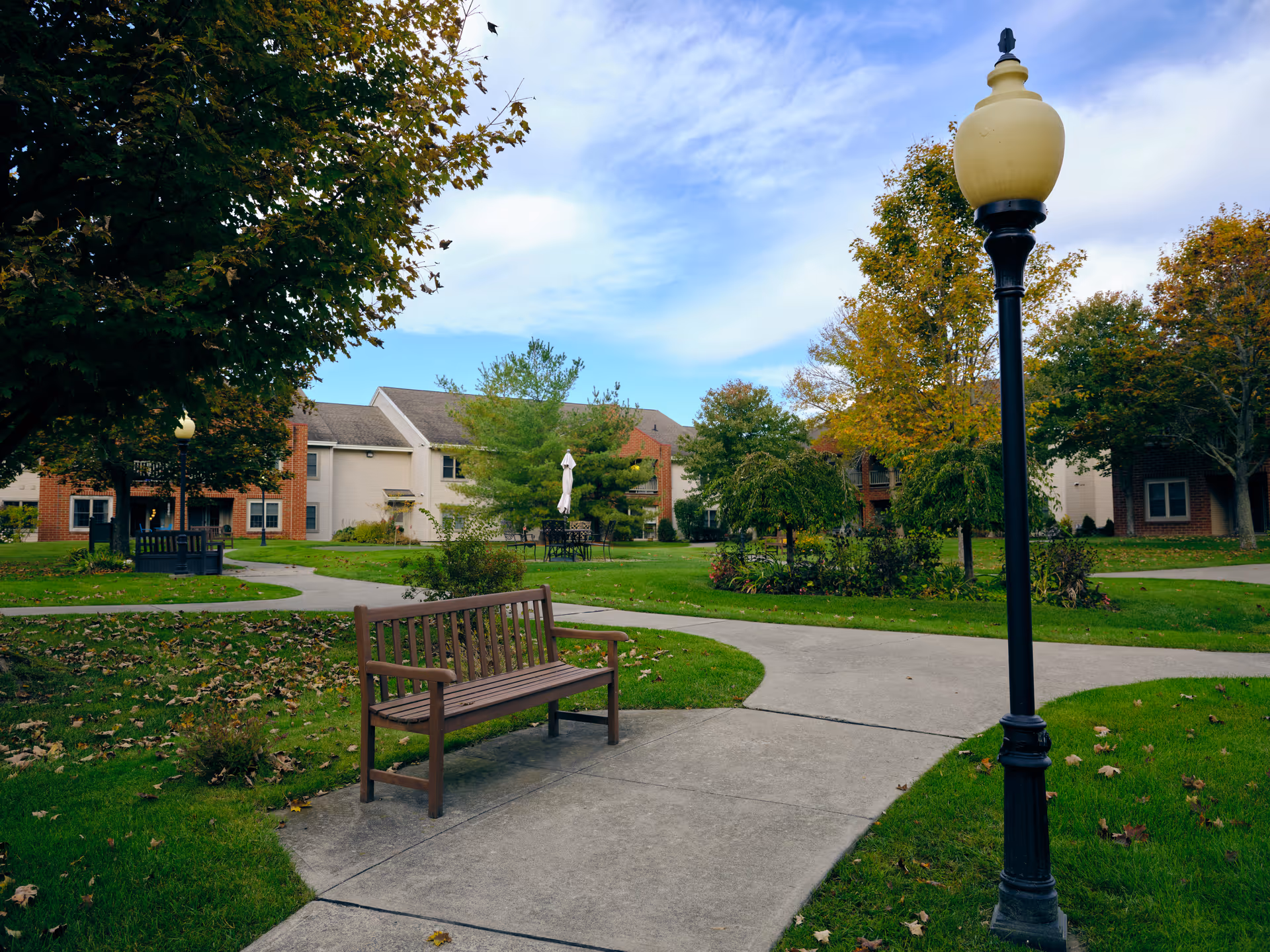 Outdoor area of a senior living facility with a paved walkway, a wooden bench, a black lamppost, green grass, trees with autumn leaves, and residential buildings in the background under a partly cloudy sky.