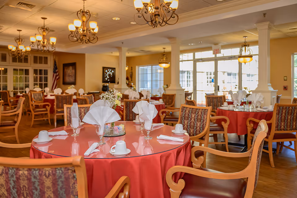 A dining room in a senior living facility with round tables covered in red tablecloths, each set with white napkins folded in decorative shapes, cups, and glassware. The room features wooden chairs with patterned upholstery, chandeliers hanging from the ceiling, and large windows letting in natural light.