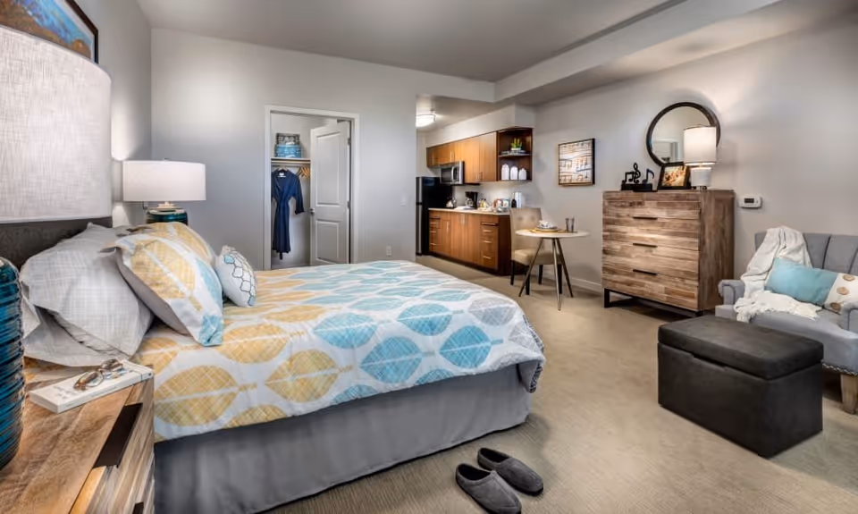 A well-lit studio apartment featuring a bed with patterned bedding in yellow and blue, a bedside table with a lamp and book, an open closet with hanging clothes, a kitchenette with wooden cabinets and appliances, a small dining table with two chairs, a wooden dresser with a round mirror and lamp, and a gray armchair with a throw blanket and pillows. A pair of slippers is placed on the carpeted floor near the bed.
