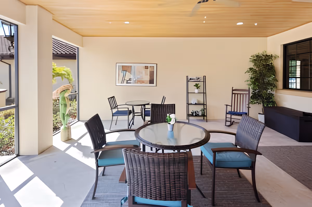 A bright indoor seating area with glass-top tables and cushioned chairs. The room has large windows letting in natural light, a wooden ceiling, a framed abstract painting on the wall, a small black shelving unit with plants and decor, a rocking chair, and a large potted plant near a window.