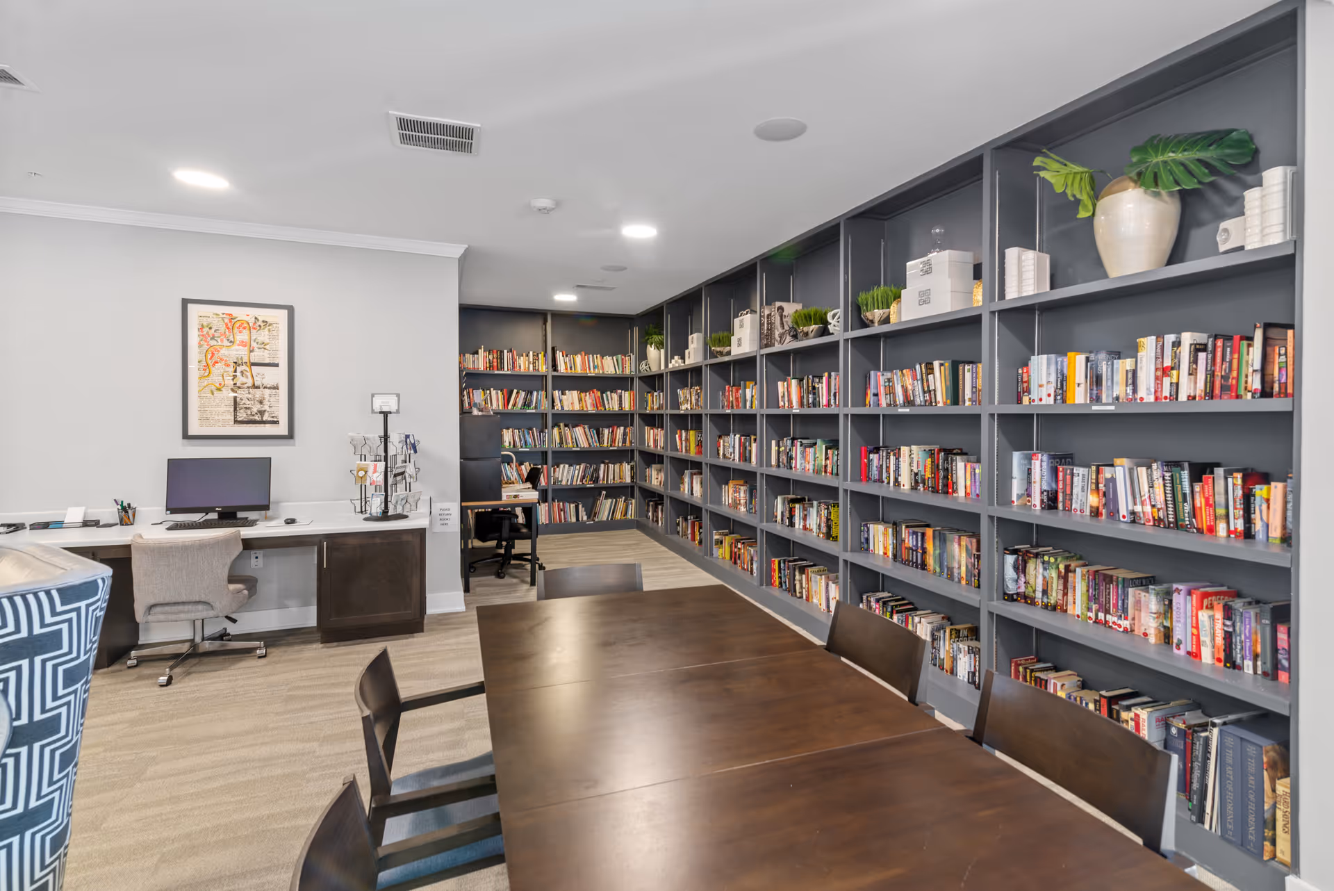 Bright communal library and activity room with a long wooden table, chairs, computer workstations and wall-to-wall bookshelves.