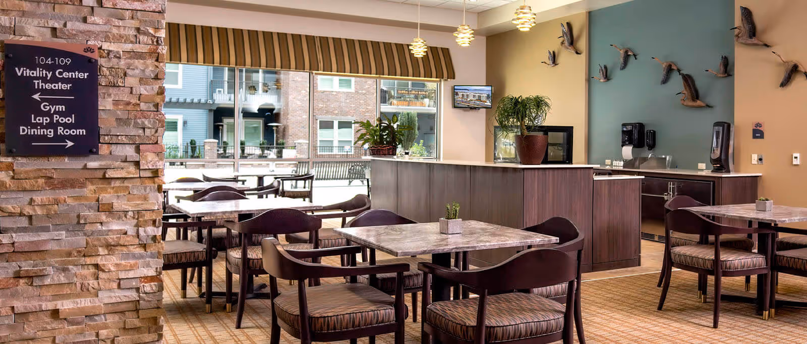 A dining area with several square tables and cushioned chairs. The room features a stone pillar with a directional sign pointing to the Vitality Center Theater, Gym, Lap Pool, and Dining Room. Large windows allow natural light to enter, showing an outdoor patio with benches. The interior has a counter with plants, a mounted TV, and decorative bird sculptures on a teal accent wall.