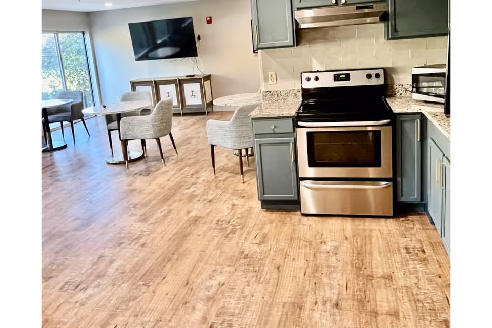 Interior view of a senior living facility's kitchen and dining area featuring a stainless steel stove, blue cabinetry, granite countertops, wooden flooring, round dining tables with gray upholstered chairs, a wall-mounted flat screen TV, and large windows letting in natural light.