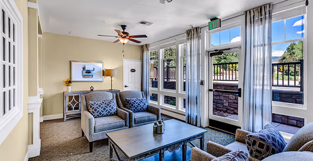 Bright common living room with upholstered armchairs, a coffee table, side table and ceiling fan next to large windows and a glass door to the outside.