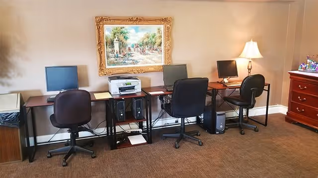 A computer workstation area with three desks, each equipped with a computer monitor and a black office chair. A printer is placed on a small table between the first and second desks. A framed painting hangs on the beige wall above the workstations. A table lamp is on the right side desk, and a wooden chest of drawers is visible on the far right.