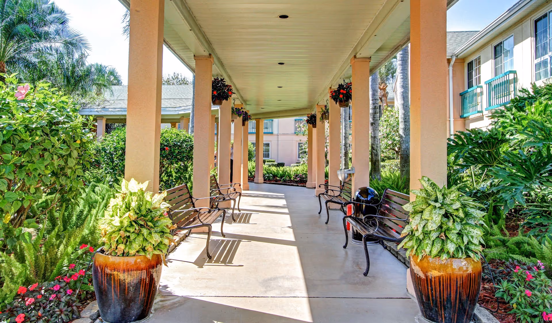 Covered outdoor walkway lined with columns, benches, hanging baskets and large potted plants at a senior living facility.