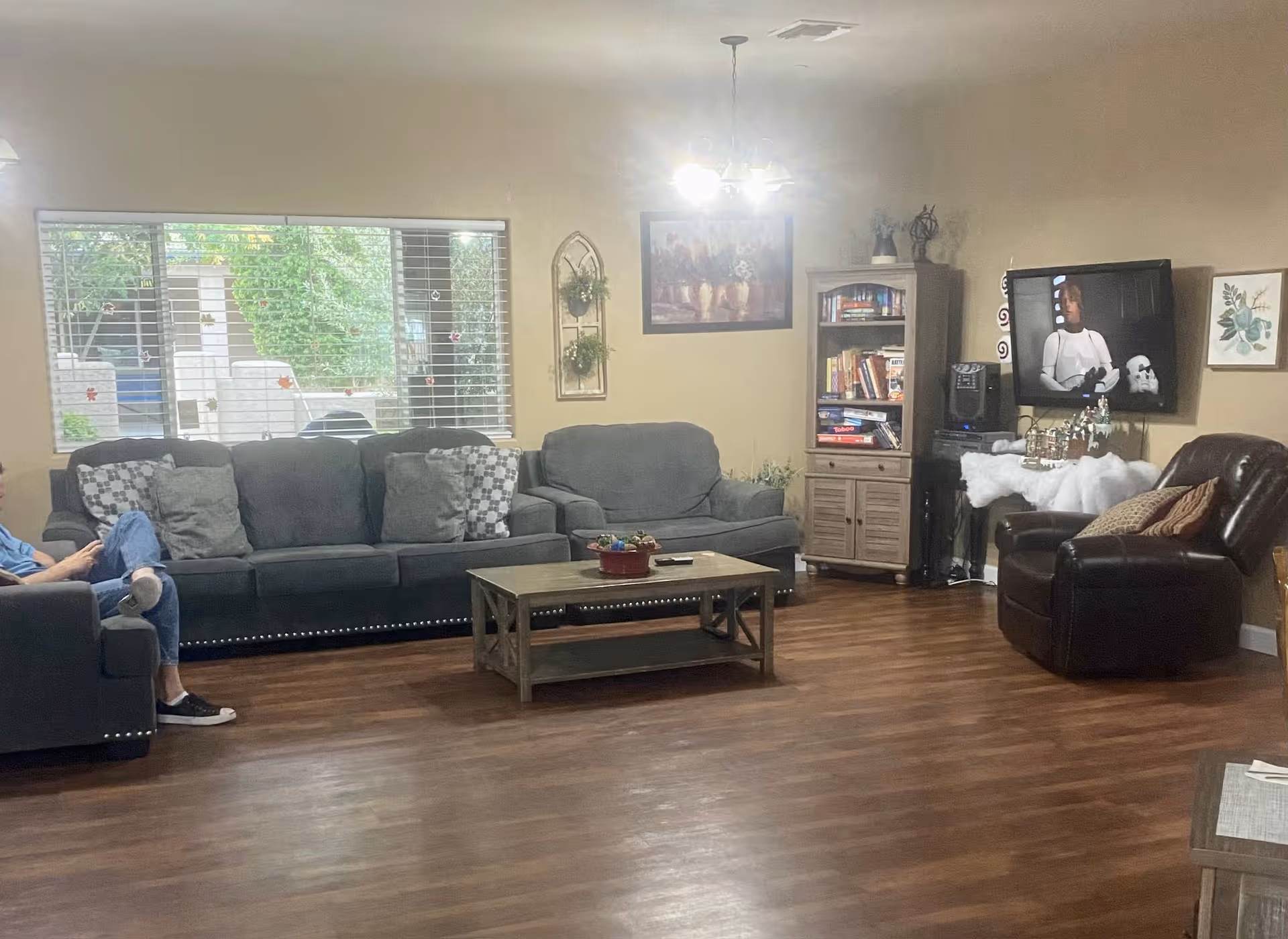 A cozy living room with a large gray sofa, a matching loveseat, and a dark brown leather recliner. A wooden coffee table sits in the center on a wood floor. A window with blinds shows greenery outside. A bookshelf filled with books and games is next to a wall-mounted TV displaying a scene from a movie. The walls are decorated with framed pictures and a hanging plant holder. A person is sitting on the left side of the sofa, partially visible.