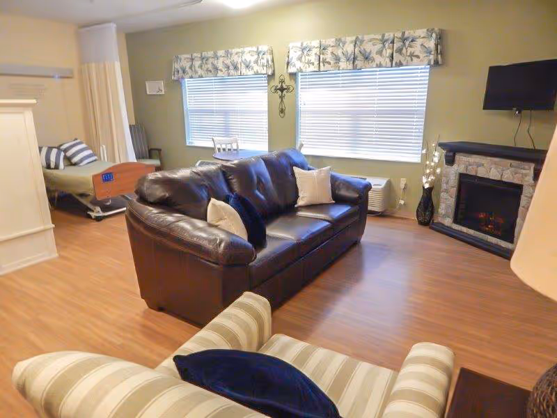 A cozy living area in a senior care facility featuring a dark brown leather sofa with light-colored pillows, a striped armchair with a dark pillow, a flat-screen TV mounted above a stone fireplace, and two windows with floral valances. In the background, there is a hospital-style bed partially separated by a curtain, indicating a combined living and sleeping space.