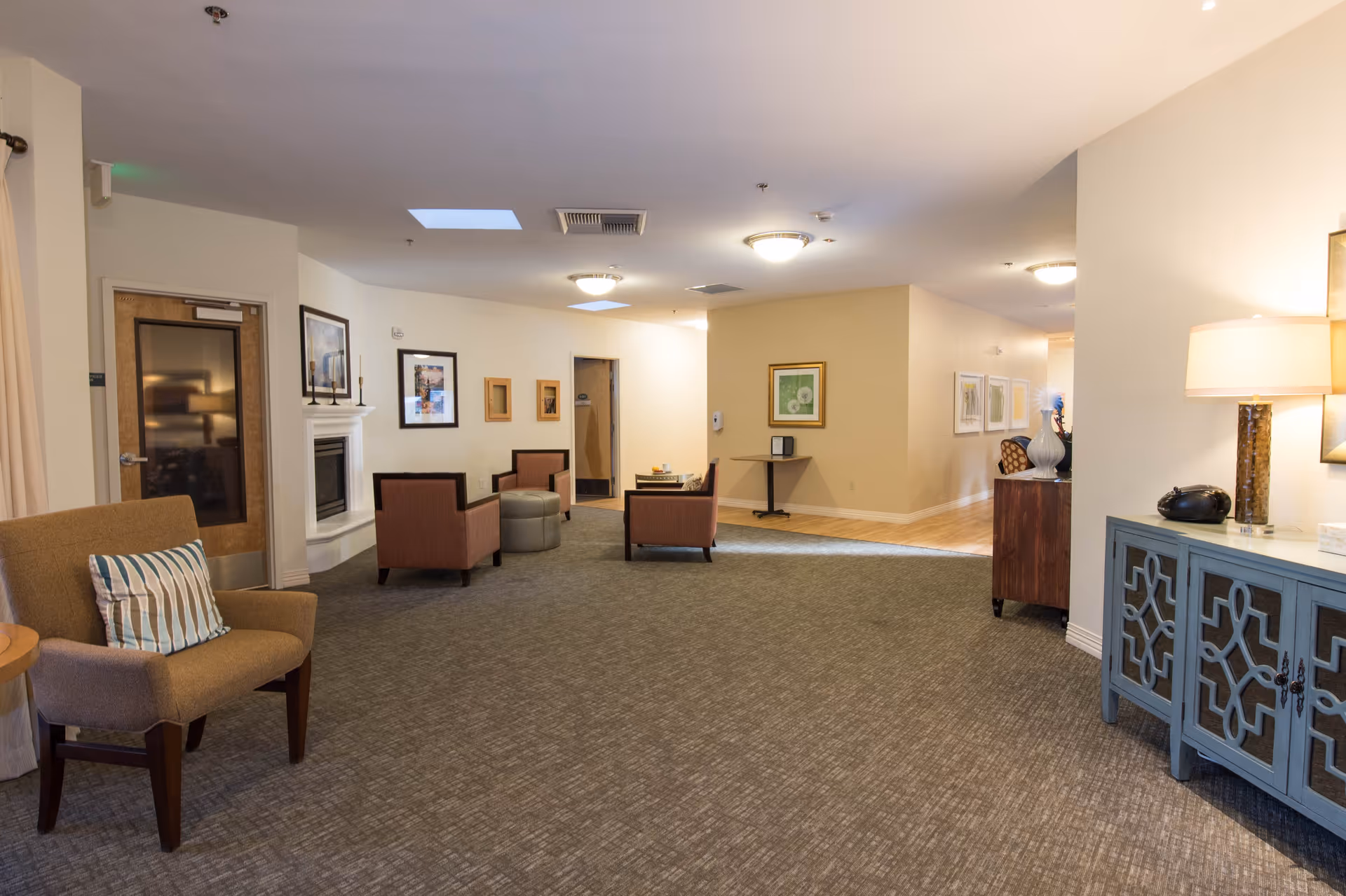 A spacious and well-lit common area in a senior living facility featuring several armchairs with cushions, a small round ottoman, framed artwork on the walls, a fireplace, and a blue cabinet with a lamp and decorative items on top. The room has carpeted flooring with a section of hardwood near the back, and ceiling lights providing warm illumination.