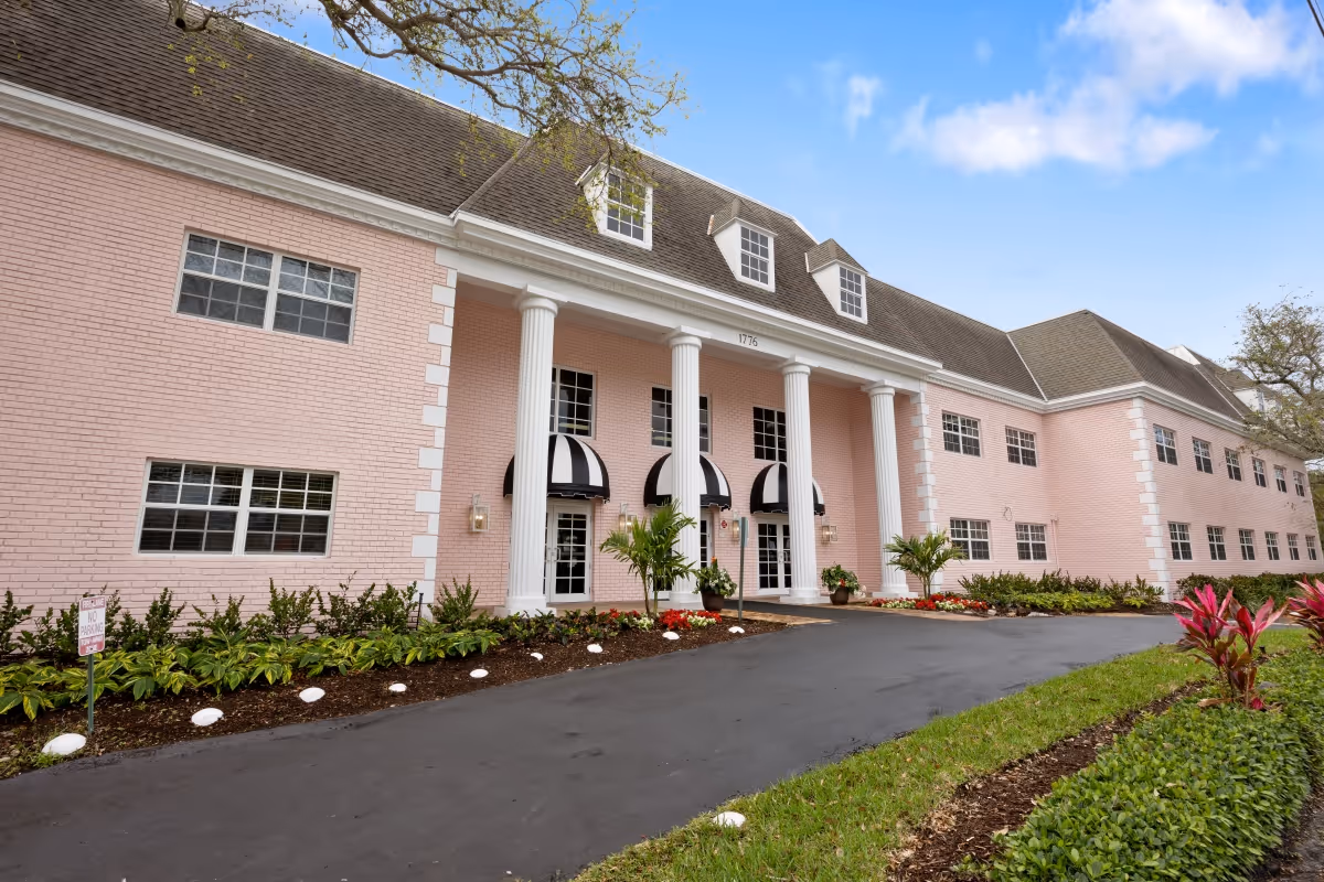 Exterior view of a large pink brick building with white columns and black-and-white striped awnings over the entrance doors. The building is surrounded by landscaped greenery and a paved driveway under a partly cloudy blue sky.