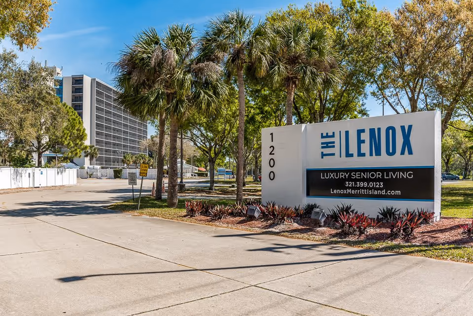 Outdoor view of The Lenox at Merritt Island senior living facility entrance sign with palm trees and a multi-story building in the background under a clear blue sky.
