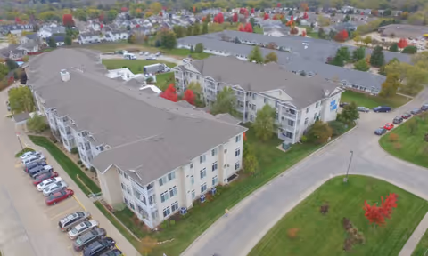 Aerial view of a multi-building senior living complex with parking, roads, and autumn trees in a surrounding neighborhood.