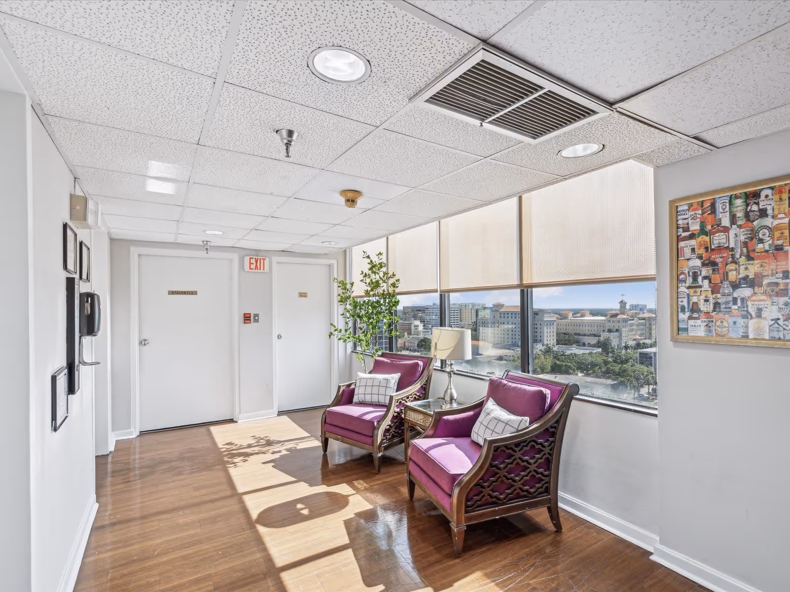 A bright hallway area with two purple cushioned wooden chairs and a small table with a lamp between them. Large windows with beige roller shades provide a view of a cityscape outside. The floor is wooden, and the ceiling has white tiles with recessed lighting and ventilation. There are two white doors labeled 'Stairwell' and 'Storage' with an exit sign above one door. A framed picture of various liquor bottles hangs on the wall.