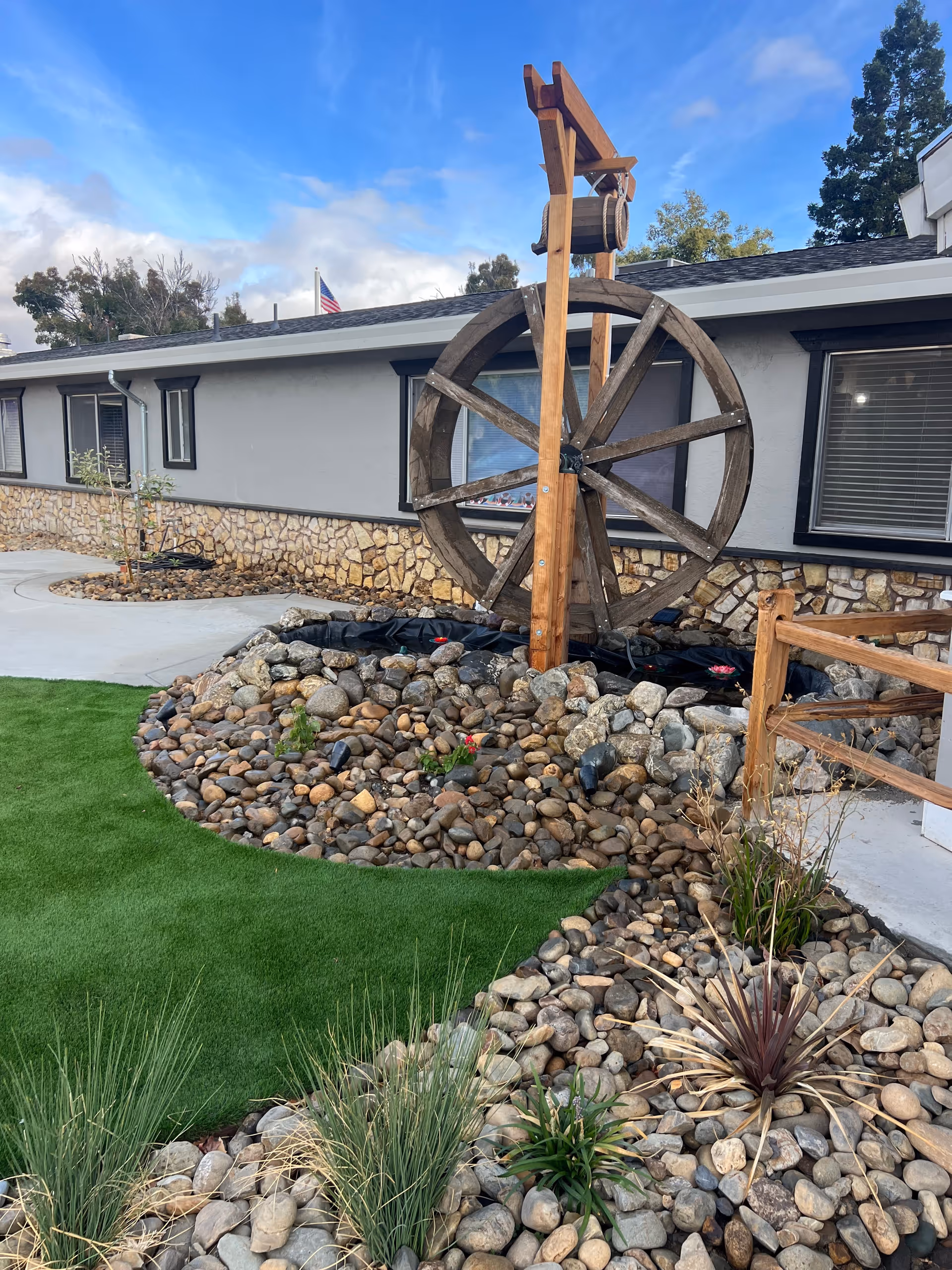 Outdoor garden area at Vacaville Ranch Post Acute featuring a large wooden water wheel surrounded by rocks and small plants, with a building in the background under a partly cloudy blue sky.