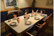 A dining room table set for six people with white tablecloth, plates, silverware, and folded peach-colored napkins. The room has wooden chairs with cushions, a side table with decorative items, and framed artwork on the walls.