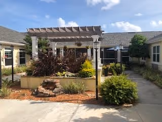 Outdoor courtyard area at The Addison of Oakleaf featuring a pergola over a raised garden bed with various plants and shrubs, surrounded by a paved walkway and building walls under a partly cloudy sky.