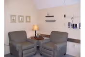 Two upholstered armchairs flank a small wooden side table with a lamp in a beige-walled sitting area decorated with framed art.
