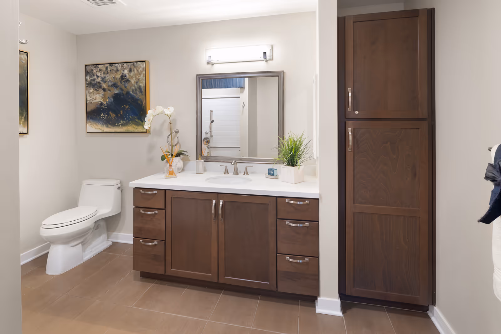 A modern bathroom featuring a white toilet, a wooden vanity with a white countertop and built-in sink, a rectangular mirror above the sink, a tall wooden cabinet to the right, and decorative items including a plant and a flower vase on the countertop. The walls are light-colored and there is a framed abstract painting on the wall above the toilet.