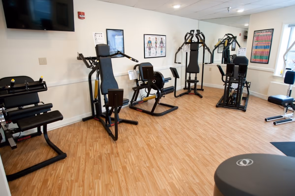 A fitness room with several exercise machines arranged along the walls. The room has light wood flooring, white walls, a large mirror, and a wall-mounted TV. Posters related to exercise and anatomy are displayed on the walls.