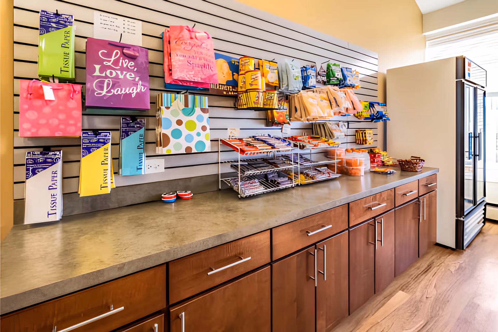 A countertop market area with hanging gift bags, snack racks, cabinets, and a glass-front refrigerator.