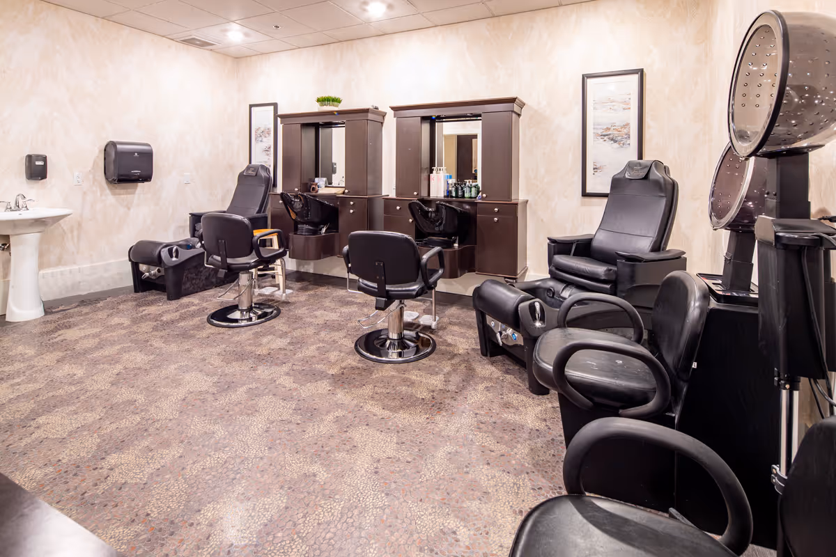 Interior view of a salon room with multiple black salon chairs, two hair washing stations with sinks, two hair drying chairs with hooded dryers, a pedestal sink, and wall-mounted cabinets with mirrors. The room has patterned flooring and light-colored walls with framed artwork.