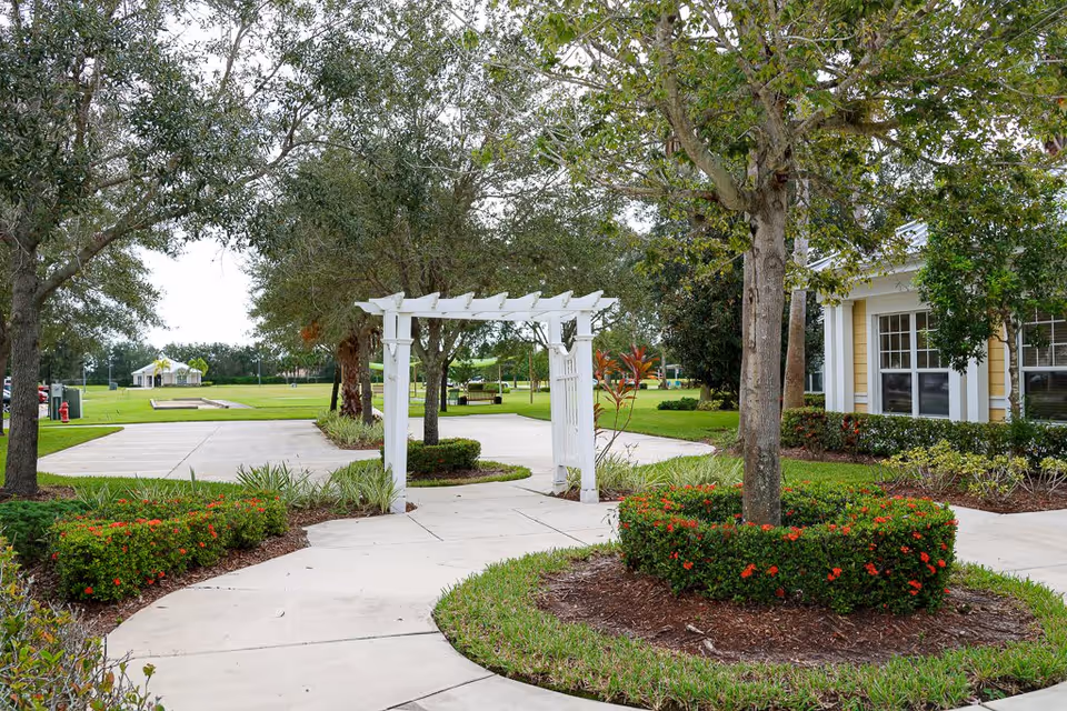 A landscaped outdoor area at The Brennity at Tradition Senior Living featuring a white wooden pergola over a paved walkway, surrounded by green grass, trees, and neatly trimmed bushes with red flowers. A yellow building with white trim is partially visible on the right side.