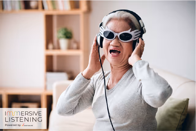 An elderly woman wearing novelty sunglasses and headphones is sitting on a couch in a living room, smiling and enjoying music. Behind her is a wooden shelf with books and a potted plant.