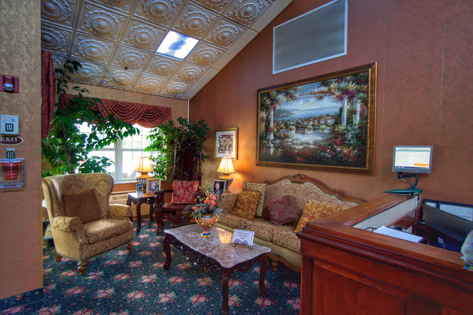 A cozy sitting area in a nursing and rehabilitation center featuring a patterned armchair and a matching sofa with decorative pillows. A wooden coffee table with a floral centerpiece and a small sign is in front of the sofa. The room has floral carpet, a large framed painting on the wall, two table lamps, and several green plants near a window with red drapes. A wooden reception desk with a computer monitor is visible on the right side.