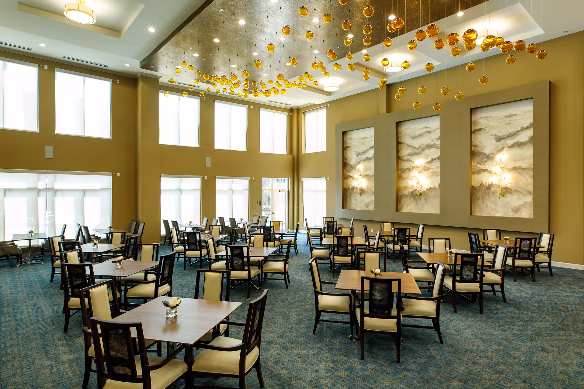 A spacious dining room with multiple square wooden tables and cushioned chairs arranged neatly. The room features large windows allowing natural light to fill the space, a high ceiling with modern light fixtures, and decorative golden hanging orbs. The walls have three large framed abstract art pieces with wall sconces.