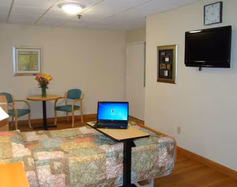 Senior living bedroom with quilted bed, laptop on table, wall TV, and small seating area.