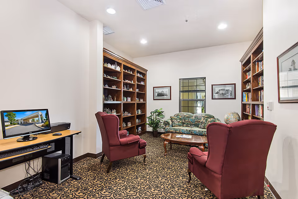 A cozy sitting area in a retirement community featuring two red upholstered armchairs, a floral patterned sofa, a wooden coffee table, and bookshelves filled with books and decorative items. A computer desk with a monitor and printer is positioned against one wall, and framed black-and-white photographs hang on the walls.