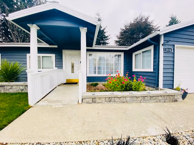 Front exterior view of a single-story blue house with white trim, a covered porch with white railings and columns, a flower bed with colorful flowers, and a driveway leading up to the entrance. The house number 2514 is visible near the garage door.