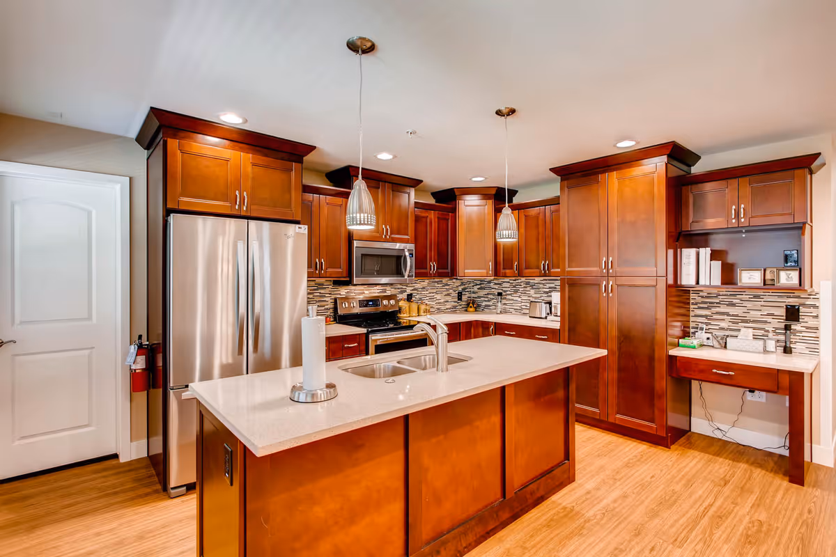 Modern kitchen with wooden cabinets, a large island with a sink and paper towel holder, stainless steel refrigerator, microwave, and stove. The backsplash features a mosaic tile design, and two pendant lights hang above the island. There is a small desk area with books and framed pictures on the right side.
