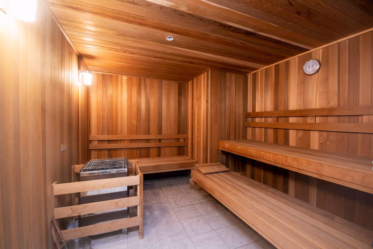 Interior of a wooden sauna room with wooden benches along two walls, a heater with stones on the left side, a light fixture on the wall, and a clock mounted on the wall.
