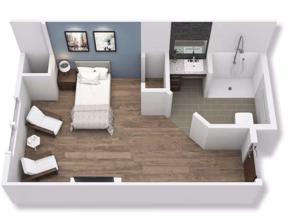 3D floor plan of a senior living facility room featuring a bedroom area with a bed, two chairs, a small table, and a nightstand with a lamp. The room has wooden flooring and two framed pictures on the wall behind the bed. Adjacent to the bedroom is a bathroom area with a sink, mirror, shower, and toilet, separated by a partial wall.