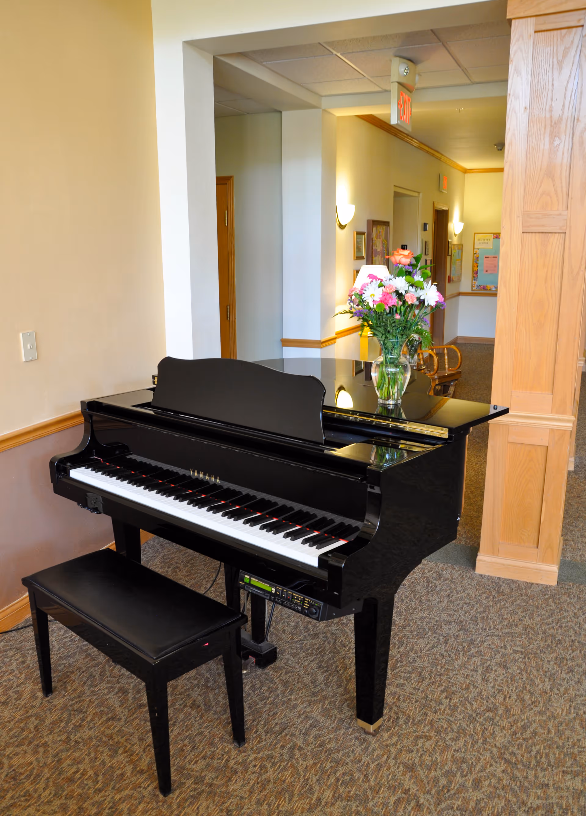 A black grand piano with a matching bench is placed in a carpeted hallway. On top of the piano is a glass vase filled with a colorful bouquet of flowers. The hallway has beige walls with wooden trim and several doors and wall-mounted lights are visible in the background.