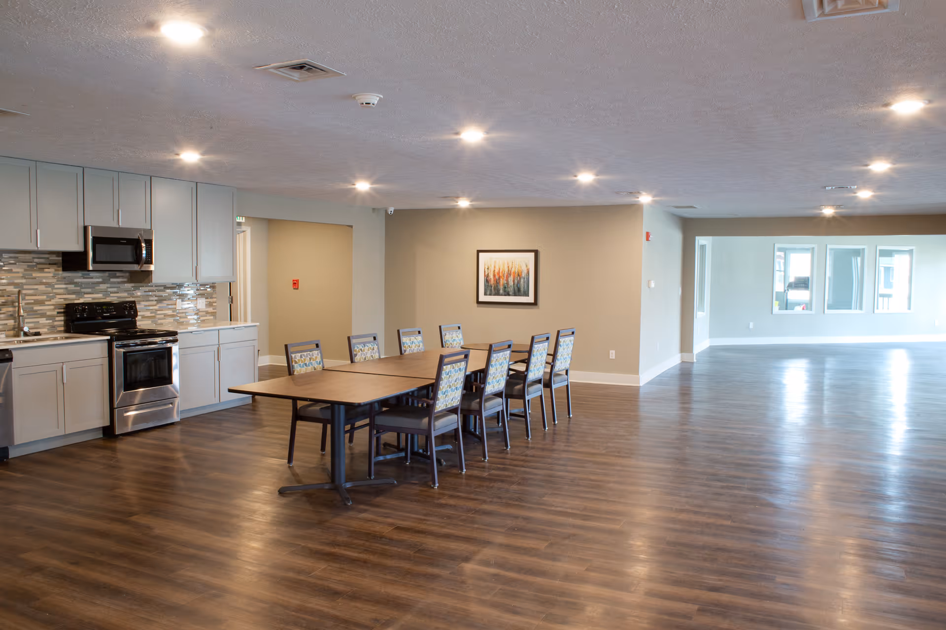 A spacious room with a modern kitchen area on the left featuring gray cabinets, a stove, microwave, and dishwasher. In the center, there is a long wooden table with eight chairs that have patterned upholstery. The room has wood flooring, beige walls, recessed ceiling lights, and a framed abstract painting on the wall. Large windows are visible in the background on the right side.