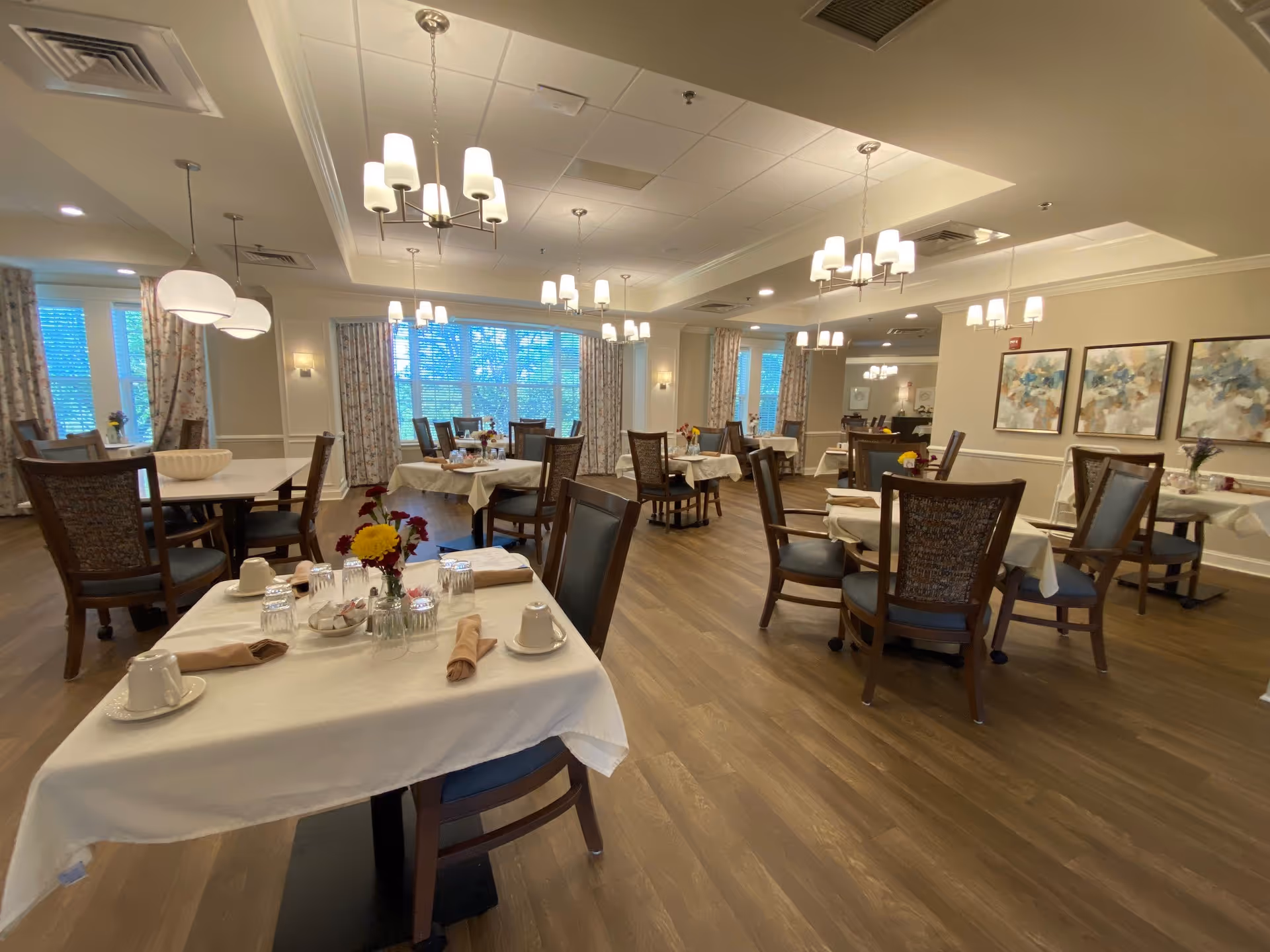 Spacious dining room with multiple set tables and chairs, chandeliers, and large windows in a senior living facility.