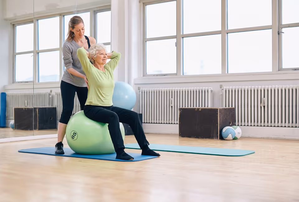 An elderly woman in a green long-sleeve shirt and black pants is sitting on a green exercise ball on a blue mat, performing a stretch with her hands behind her head. A younger woman in a gray top and black pants is standing behind her, assisting with the stretch in a bright exercise room with large windows and wooden floors.