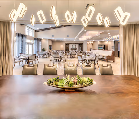 A spacious and well-lit dining room with multiple tables and chairs arranged neatly. Modern pendant lights hang from the ceiling, and large windows with curtains allow natural light to fill the room. A decorative centerpiece with succulents is placed on a wooden table in the foreground.