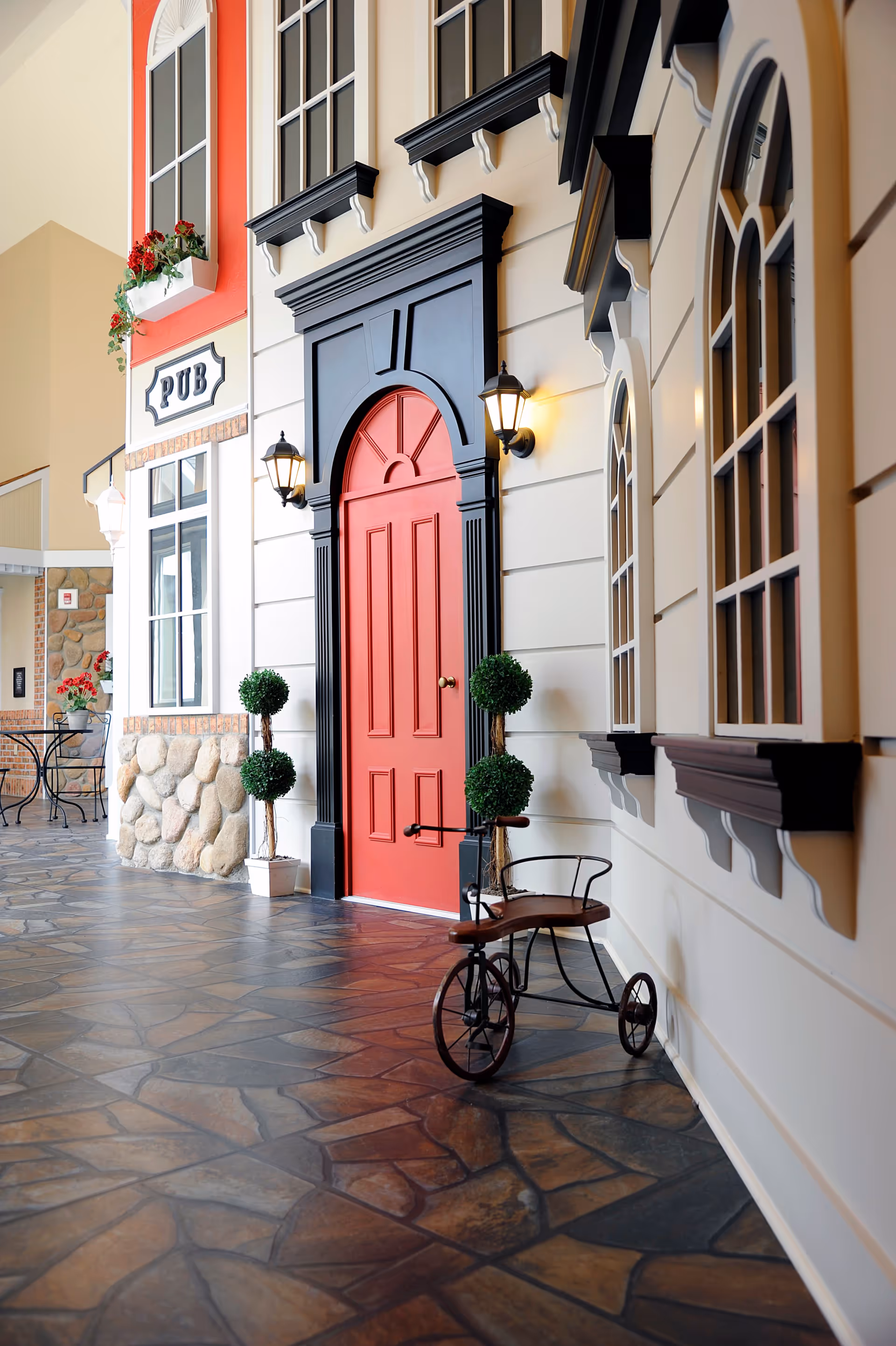 Indoor corridor styled like a small village street with a red door framed in black, wall lanterns, potted topiaries, a small tricycle, and a 'PUB' storefront sign.