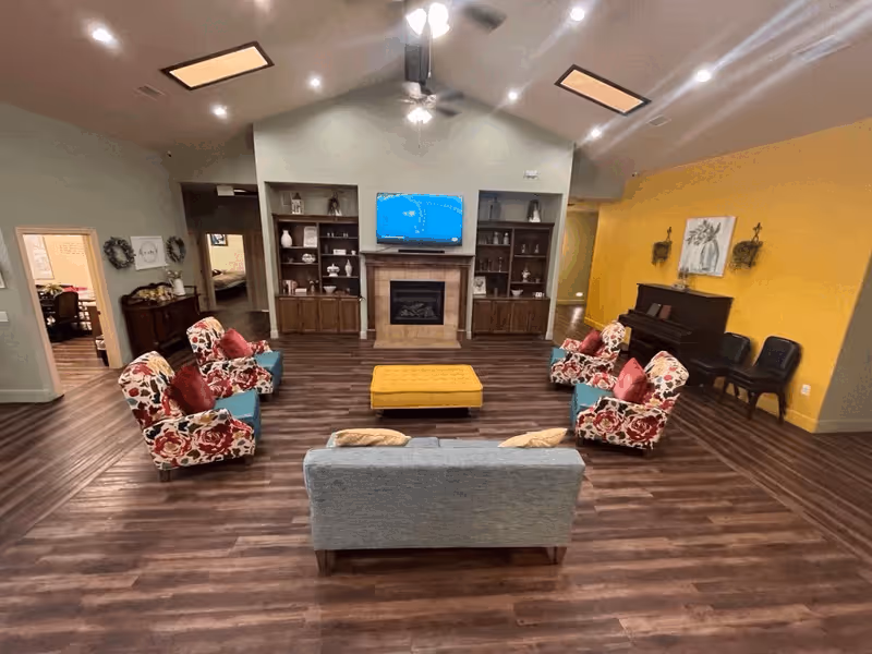 A spacious senior living common area with wood flooring, a gray couch, four floral armchairs with red cushions, and a yellow ottoman in the center. The room features a fireplace with a TV mounted above it, built-in wooden shelves on either side, and a piano against a yellow accent wall. The ceiling is vaulted with recessed lighting and a ceiling fan.