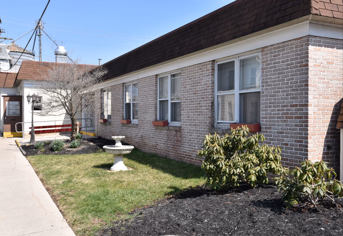 Low brick building with several windows, a small lawn featuring a white birdbath and shrubs, and a sidewalk leading to an entrance.