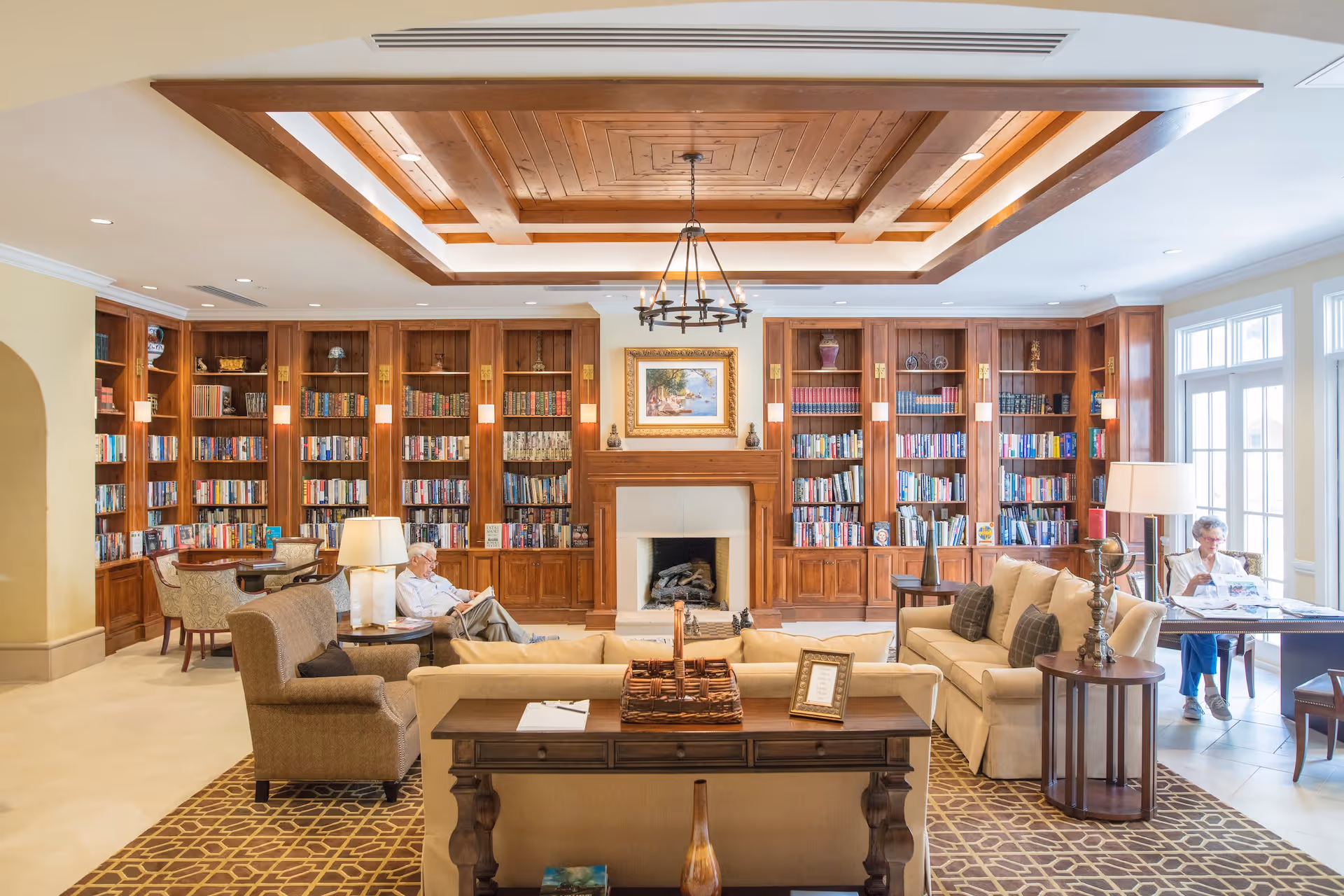 A bright and spacious library room with wooden bookshelves filled with books lining the back wall. There is a fireplace in the center with a framed painting above it. Comfortable seating including armchairs and sofas are arranged around a wooden table with a basket and picture frame. Two elderly people are seated, one reading a book and the other reading a newspaper near large windows letting in natural light. The ceiling features wooden beams and a chandelier.