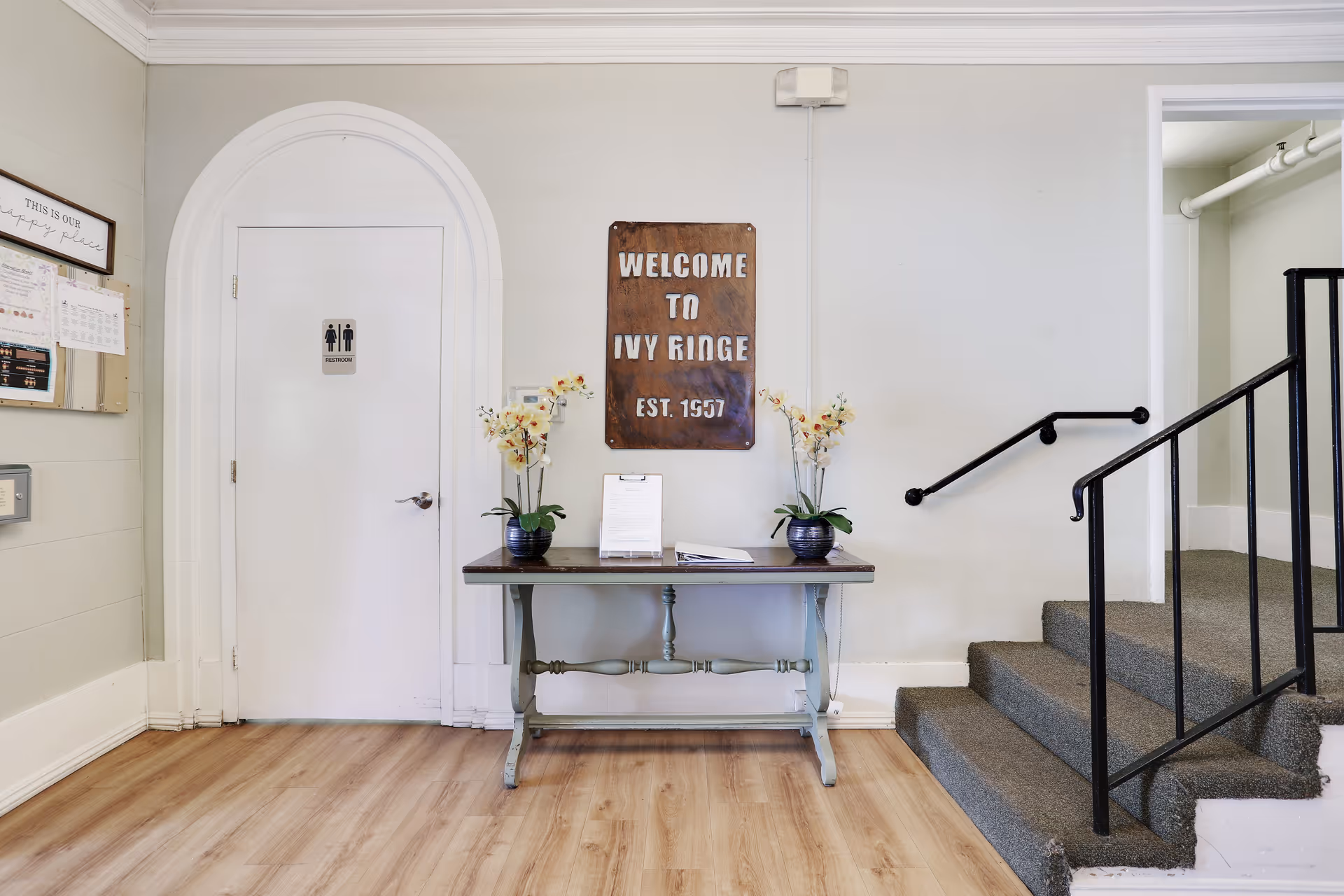 Entry foyer with a welcome sign for Ivy Ridge above a table with flowers, a restroom door on the left, and carpeted stairs to the right.