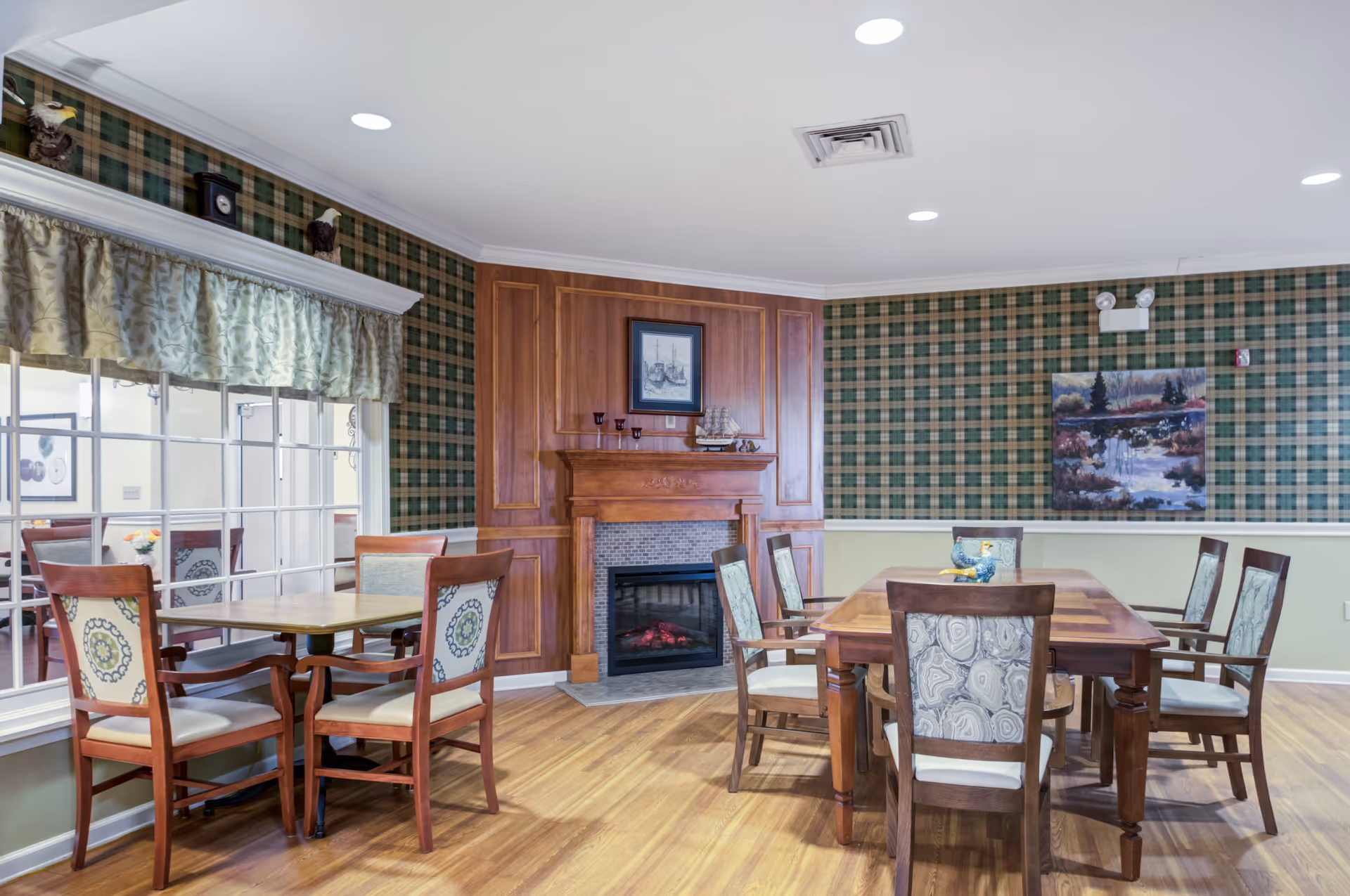 A cozy dining area with wooden tables and chairs featuring patterned upholstery. The room has a wooden floor, green plaid wallpaper, and a wooden fireplace with a framed picture and decorative items on the mantel. A large window with a floral valance looks into another room.
