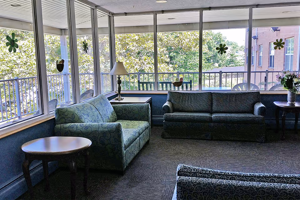 A sunlit sitting area with large windows overlooking trees and a building. The room contains two patterned sofas, a small round wooden table, a side table with a lamp, and another side table with a vase of flowers. Decorative flower-shaped window clings are on the glass.