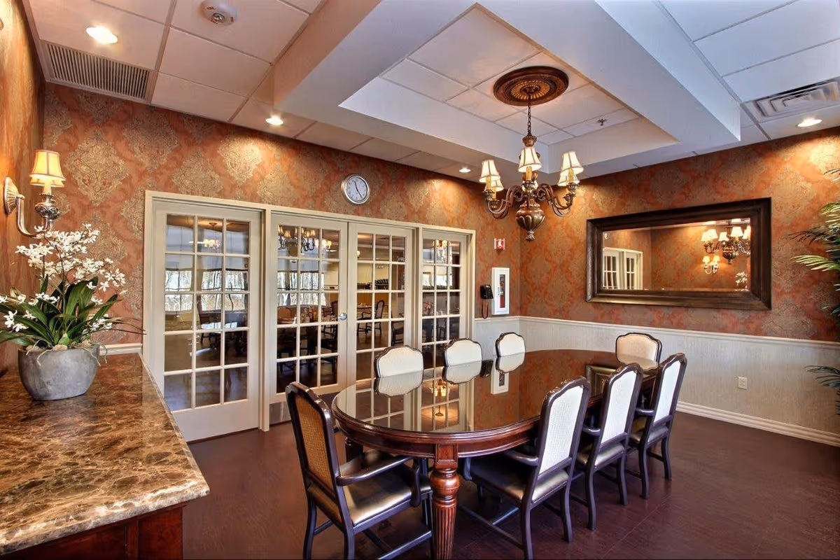A formal dining room with a large oval wooden table surrounded by eight upholstered chairs. The room features ornate wallpaper with a damask pattern, a large rectangular mirror on one wall, a chandelier hanging from the ceiling, and a marble countertop with a potted plant. French doors with glass panes lead to another room.