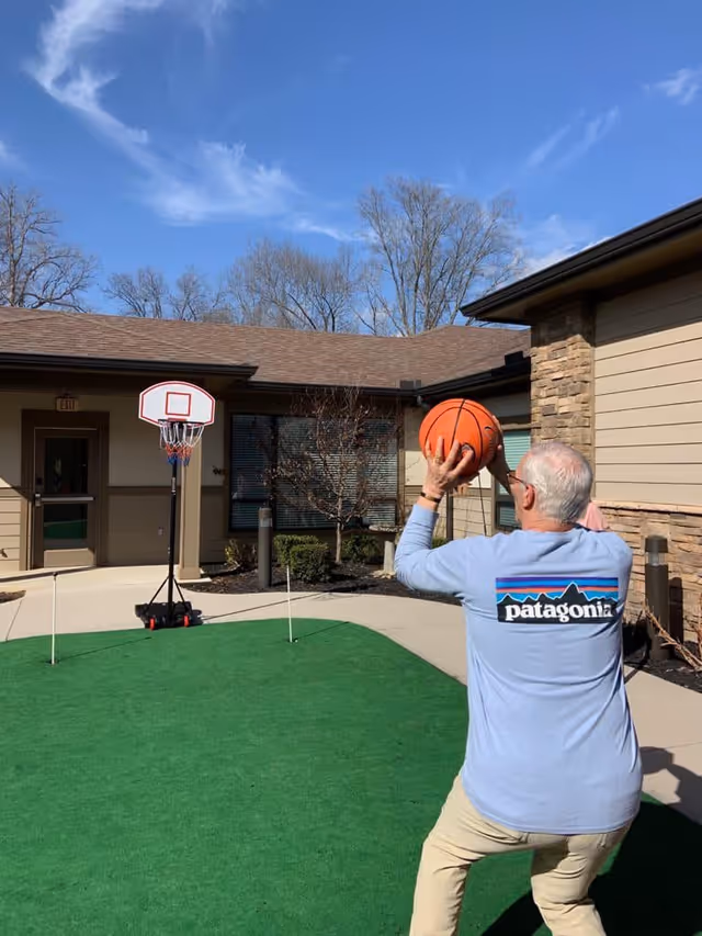 An elderly man wearing a light blue Patagonia shirt is preparing to shoot a basketball towards a small basketball hoop set up outdoors on a green artificial turf area. The scene is set in a courtyard with beige buildings and leafless trees under a clear blue sky.